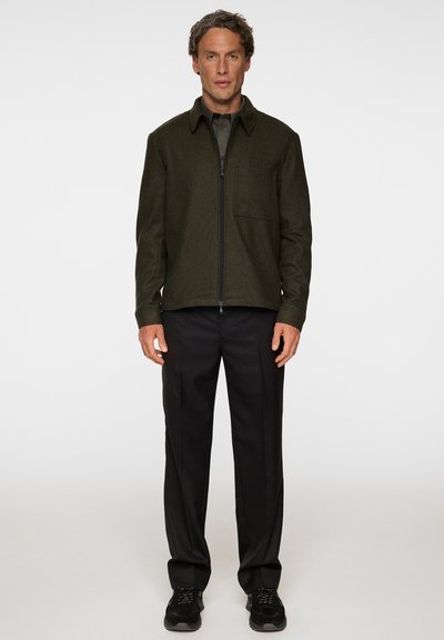 Olivgrön zip-up jacka med krage och bröstficka, matchad med svarta byxor och svarta sneakers. Texturerad, mjuk tyg.