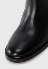 Bottillon en cuir noir avec une finition lisse, bout arrondi et coutures ton sur ton le long du bord de la semelle. Design élégant et minimaliste.