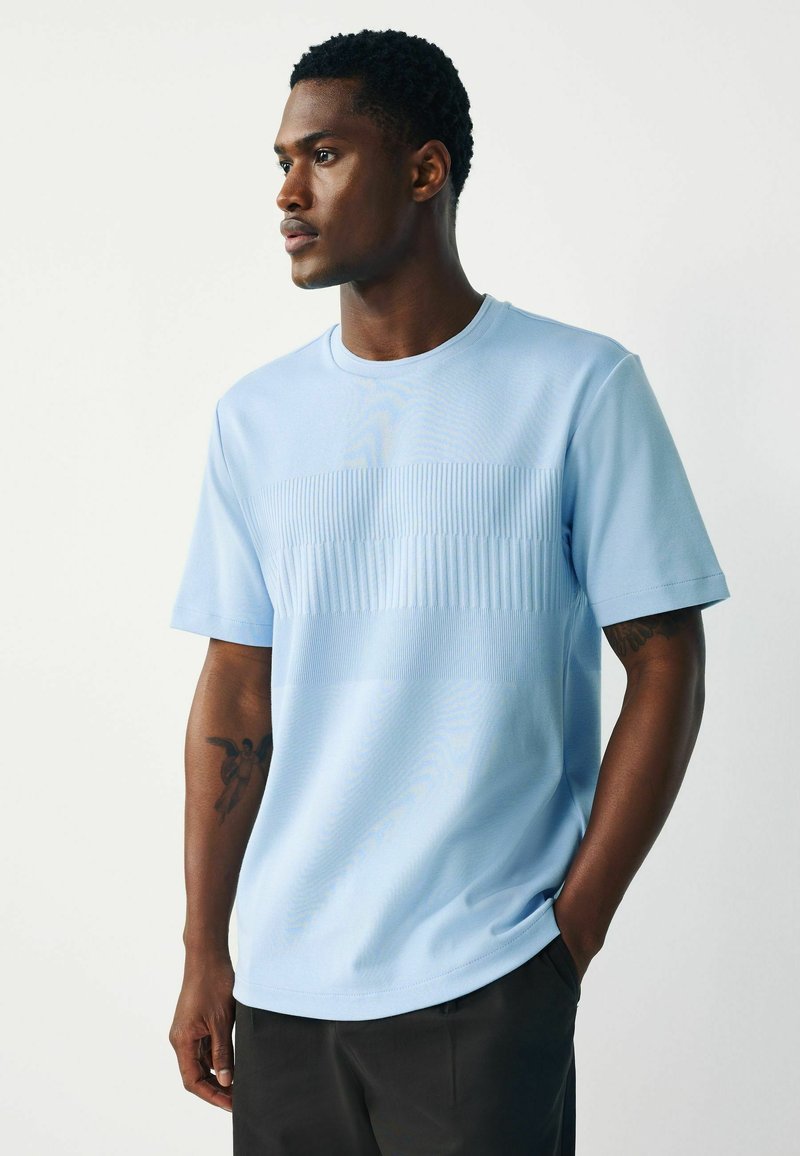 Bărbat privind în lateral, purtând o cămașă cu mâneci scurte texturată de culoare albastru deschis și pantaloni închiși la culoare, cu un tatuaj vizibil pe brațul stâng.