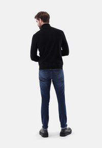 Schwarzer Strickpullover mit hohem Kragen, kombiniert mit blauen Jeans und schwarzem Schuhwerk, von hinten vor einem neutralen Hintergrund gesehen.
