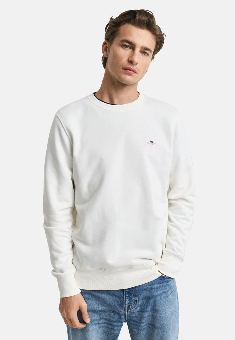Weißes Sweatshirt mit geripptem Rundhalsausschnitt und Bündchen, ausgestattet mit einem kleinen gestickten Logo auf der Brust. Kombiniert mit blauen Jeans.