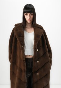Giovane donna con capelli scuri e lisci, che indossa un ampio cappotto di pelliccia marrone, un crop top bianco e pantaloni scuri, in piedi davanti a uno sfondo bianco.