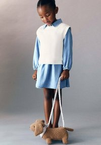 Dítě má na sobě bílou pletenou vestu přes světle modré džíny, drží zapletený vodítko připojené k plyšové hnědé psí hračce.