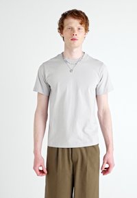 Lys grå t-shirt lavet af blødt stof, kortærmede, rund hals, tætsiddende pasform. Båret med løstsiddende olivengrønne bukser og en sølvkæde.