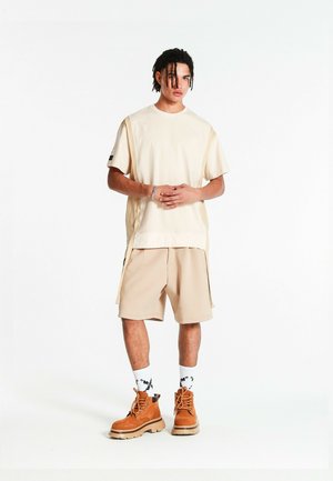 Joven de pie con las manos entrelazadas, vistiendo una camiseta beige extragrande, pantalones cortos a juego, calcetines blancos con estampado y botas de trabajo marrones sobre fondo blanco.