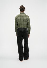 Bărbat întors cu spatele, purtând o cămașă verde cu carouri, cu mânecă lungă, băgată în jeans negri lărgi, cu adidași, pe un fundal alb simplu.