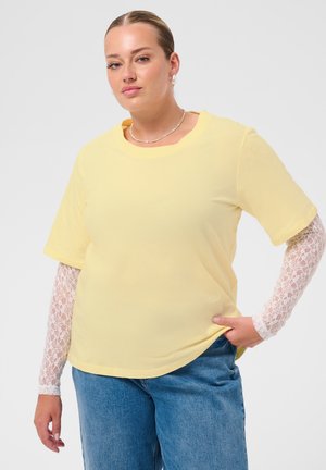 Frau mit nach hinten gegelten Haaren, die ein gelbes kurzärmliges Oberteil über weißen Spitzenärmeln und blaue Jeans trägt, steht mit einer Hand in der Tasche.