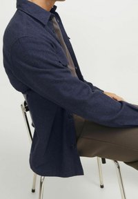 Námořnická modrá košile na knoflíky z texturované tkaniny, s špičatým límcem a dlouhými rukávy, nošená přes šedý top, s uvolněným střihem.