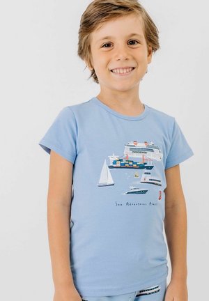 Lächelnder Junge, der ein hellblaues T-Shirt mit bunten Illustrationen von Kreuzfahrtschiffen, Frachtschiffen, Segelbooten und Schnellbooten trägt, auf dem der Text "Sea Adventures Await!" steht.