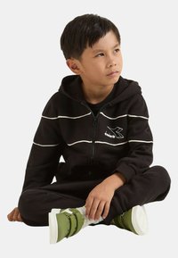 Felpa con cappuccio nera con cerniera e strisce bianche, abbinata a pantaloni coordinati. Indossando scarpe da ginnastica verdi con doppie fibbie e suole bianche.