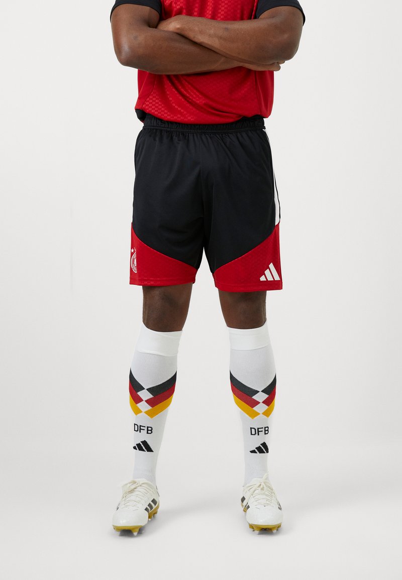 Μαύρο και κόκκινο ποδοσφαιρικό σορτσάκι με υφή πλέγματος, επισημασμένο με λευκή ρίγα. Λευκές κάλτσες μέχρι το γόνατο με σχέδια διαμαντίων σε μαύρο, κόκκινο και κίτρινο.