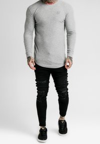 Slim-Fit langärmliges graues Ripp-Shirt mit abgerundetem Saum, kombiniert mit schwarzen, lässigen Skinny-Jeans und schwarzen Sneakers mit goldenen Akzenten.