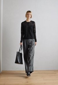 Čierny čipkovaný top s dlhým rukávom a geometrickými vzormi, v kombinácii s voľnými šedými nohavicami. Model nesie čiernu koženú tašku a má na sebe podpätky.