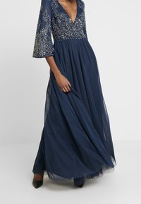 Vestido maxi azul marino con un escote en V profundo, adornado con intrincados bordados de cuentas. Presenta mangas acampanadas y transparentes, y una falda fluida.