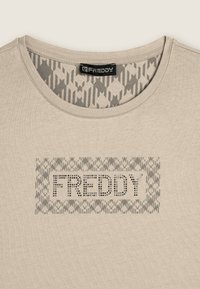 Camiseta de algodón beige con un escote redondeado, que presenta un gráfico estampado en gris y el texto "FREDDY" con tachuelas en el centro. Textura suave.