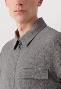 Chaqueta gris con cremallera y superficie texturizada, presenta un cuello extendido y un bolsillo en el pecho. La tela tiene un patrón suave y ligeramente elevado.