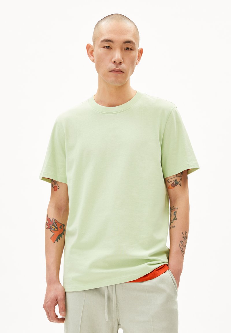 Hellgrünes T-Shirt mit kurzen Ärmeln und Rundhalsausschnitt. Das Material wirkt weich und hat eine glatte Textur. Auffällige Tattoos an den Armen mit bunten Motiven.