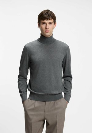 Ung mand iført en grå rullekrave og beige bukser, stående med hænderne i lommerne mod en ensfarvet hvid baggrund.