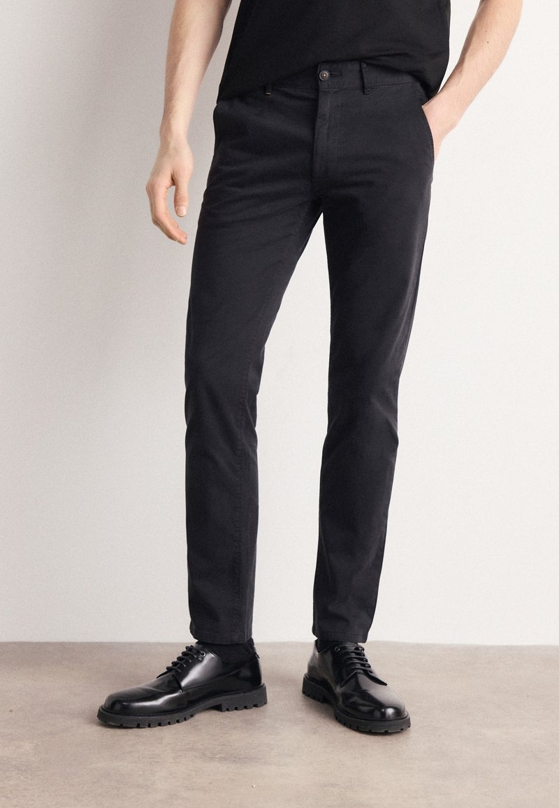 Homme portant un pantalon noir ajusté, des chaussettes noires et des chaussures habillées noires vernies à lacets, debout sur un sol gris contre un mur clair.