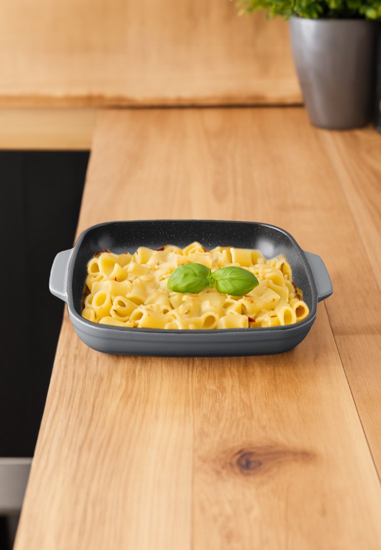 Graue rechteckige Auflaufform, gefüllt mit Pasta, garniert mit zwei Basilikumblättern, auf einer Holzoberfläche platziert.