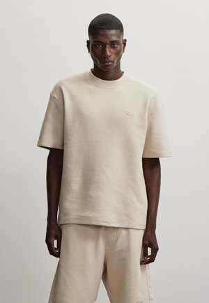 Young man with short hair wearing textured beige short-sleeve shirt and matching beige shorts, standing against a plain light background.