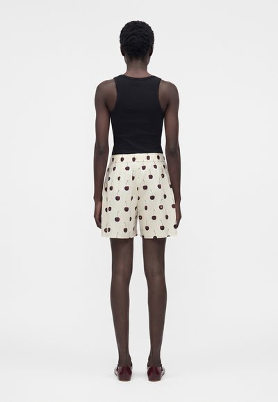 Άτομο που στέκεται με την πλάτη, φοράει μαύρο αμάνικο τοπ, λευκά σορτς με σχέδιο κερασιών και σκούρα κόκκινα παπούτσια σε απλό φόντο.