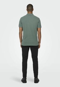 Grünes Polohemd mit kontrastierndem Kragen und Ärmelbesatz, kombiniert mit schwarzen Hosen und schwarzen Schuhen, von hinten betrachtet.