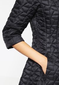 Chaqueta negra acolchada con mangas de tres cuartos, mostrando una mano metida en un bolsillo frontal sobre un fondo blanco liso.