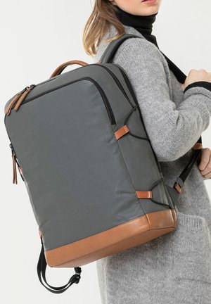 Grauer Rucksack mit glatter Außenfläche, braunen Lederakzenten, schwarzen Reißverschlüssen und verstellbaren Trägern, vor einem neutralen Hintergrund präsentiert.