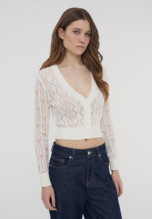 Cardigan bianco corto con scollo a V e chiusura a bottoni. Presenta intricati motivi di maglia simili al pizzo e maniche lunghe con polsini a costine.