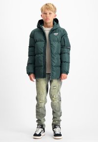Groene pufferjack met een capuchon en rits, gecombineerd met versleten lichtblauwe jeans en zwarte sneakers met witte accenten.
