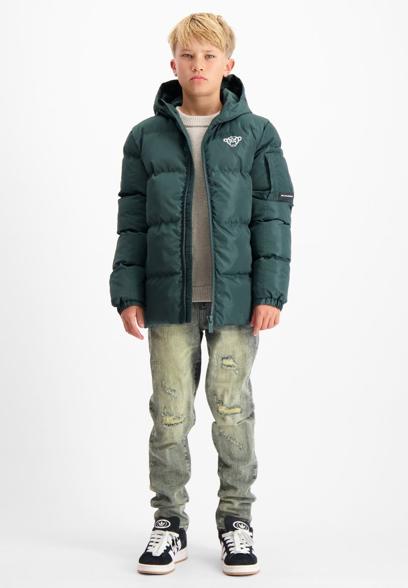 Groene pufferjack met een capuchon en rits, gecombineerd met versleten lichtblauwe jeans en zwarte sneakers met witte accenten.