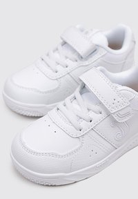 Zapatillas blancas con parte superior de cuero liso, puntera redondeada, cierre de cordones y una tira de velcro. Suela con textura antideslizante y detalle de logo lateral.