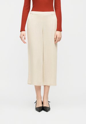 Woman wearing cream wide-leg cropped pants, rust long-sleeve top, and black pointed-toe heels, standing on a white background.