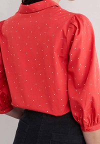 Camicia rossa con colletto, maniche corte e puff, e pois bianchi. Tessuto morbido con una texture liscia. Presenta dettagli con bottoni sui polsini delle maniche.