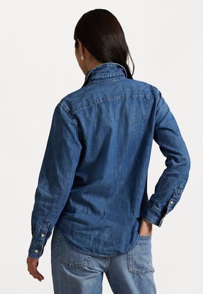 Camisa de denim leve em azul escuro, com mangas longas, colarinho, punhos com botões e um corte descontraído. Apresenta uma textura lisa.