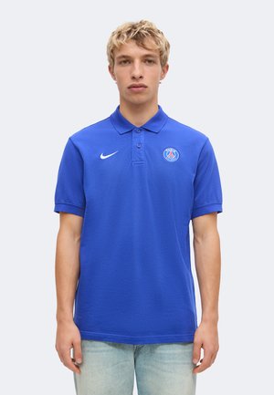 Mladý muž oblečený v modrej Nike polo košeli s logom Paris Saint-Germain stojí pred jednoduchým svetlošedým pozadím.