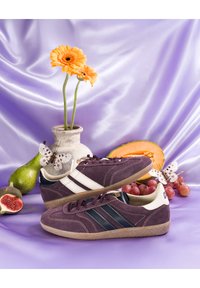Deux baskets violettes avec des rayures blanches sur satin violet, à côté d'un vase blanc avec des fleurs orange, des fruits et deux papillons blancs.