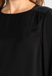 Mustavartalo paita, jossa on pyöreä pääntie, sileä pinta ja rypytetty yksityiskohta olkasaumoissa. Pitkät hihat rentoissa mitoituksissa.