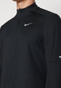 Schwarzes, langärmliges Sportoberteil mit hohem Kragen und Viertelreißverschluss. Verfügt über ein reflektierendes Nike-Logo auf der Brust. Glatte Stoffoberfläche.