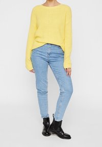 Gul stickad tröja med ribbad struktur, som kombineras med ljusblå slim fit jeans. Modellen har på sig svarta ankle boots.