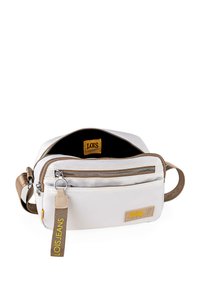 Bolso cruzado de tela blanca con acentos en color marrón claro, que incluye un bolsillo con cremallera, correa ajustable y una etiqueta de logotipo amarilla, diseño minimalista.