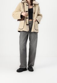 Beige sherpa-takki, jossa on ruskeat somisteet, suuret napit ja taskut. Yhdistetty harmaisiin raidallisiin housuihin ja mustiin paksupohjaisiin kenkiin. Ruskea käsilaukku.