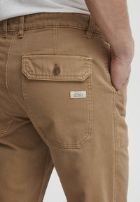 Brown cotton pants with a textured finish, featuring a buttoned back pocket and a small fabric label on the right pocket.