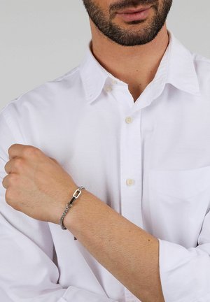 Braccialetto a catena d'argento con una chiusura rettangolare. Indossato su un polso nudo, abbinato a una camicia bianca con colletto. Texture liscia.