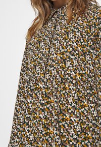 Femme portant une robe à manches longues avec un col montant et un petit imprimé floral aux tons blanc, jaune, vert et marron.