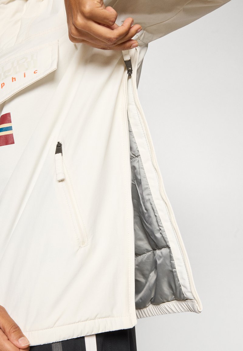 Chaqueta ligera de crema con un bolsillo con cremallera, forro gris y detalle de rayas. Presenta una tela texturizada y un diseño de corte relajado.