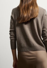 Suéter de punto marrón con mangas largas y un dobladillo acanalado. El material parece suave y texturizado, combinado con una falda beige lisa.