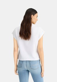 T-shirt blanc à manches courtes avec une coupe décontractée, présentant un col rond et une texture lisse, associé à un jean bleu clair moulant.