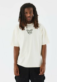 Camiseta de gran tamaño en color crema con un gráfico de mariposa en verde oliva en el pecho. Mangas cortas y un ajuste relajado. Textura suave.